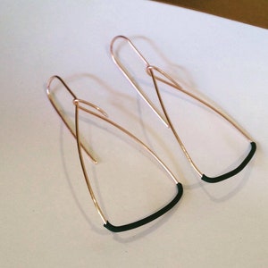 May include: A pair of gold-colored wire earrings with a teardrop shape. The bottom of each earring features a curved, dark green accent. The earrings are displayed on a white surface.