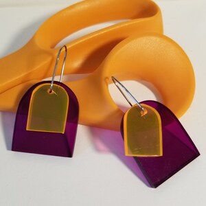 May include: A pair of modern earrings with a geometric design. Each earring features a translucent purple arch shape with a smaller, yellow-orange arch layered on top. The earrings hang from silver-colored ear wires. A pair of orange scissors is in the background.