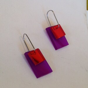 May include: A pair of modern earrings featuring a rectangular purple base with a smaller red square accent. The earrings have silver-colored, curved ear wires. The design is minimalist and geometric.