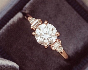 May include: A rose gold engagement ring with a large, round, clear center stone and smaller, rectangular accent stones. The ring is set in a dark blue velvet ring box.