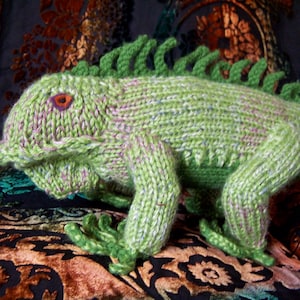 May include: A green knitted iguana with pink accents. The iguana is sitting on a patterned fabric.