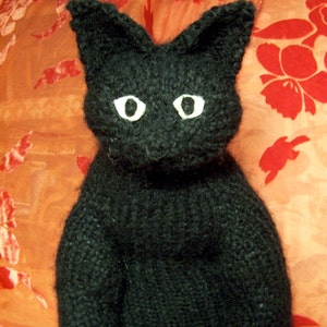 May include: A black knitted cat toy with white eyes. The cat is sitting on a red and orange patterned fabric.