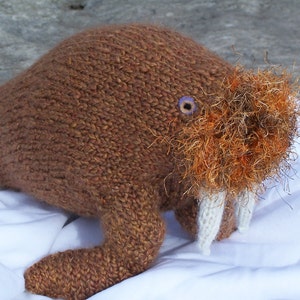 May include: A brown knitted walrus with a white tusk, orange fur, and a purple eye. The walrus is lying on a white surface.