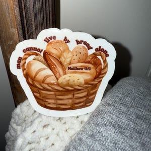May include: A sticker featuring a watercolor illustration of a basket filled with various types of bread. The sticker has the text "Man cannot live by bread alone" and "Matthew 4:4" in a brown font.
