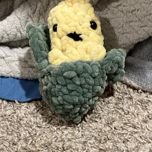 May include: A crocheted corn on the cob plush toy with a green husk and yellow kernels. The toy has black eyes and a black mouth.