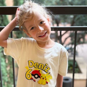 May include: A cream-colored t-shirt featuring a graphic of Pikachu and a Poké Ball, with the name "Deniz" and the number "6" in yellow and red. The child is smiling and has their hand in their hair.