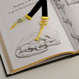 May include: Black velvet bookmark with gold-colored accents and a key charm. The bookmark features a feather charm and green beads. The open book displays a dragon illustration and the text "Reading is the quietest to go on the grandest ad".