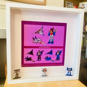 May include: A framed print of Transformers characters Starscream and Thundercracker in their jet forms. The print is on a pink background with glitter. The frame is white and has a white mat. There are also small figurines of Starscream and Thundercracker in their robot forms.