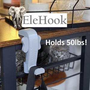 May include: A gray plastic hook shaped like an elephant with the text "EleHook" and "Holds 50lbs!"  The hook is attached to a wooden table and a black strap is hanging from it.