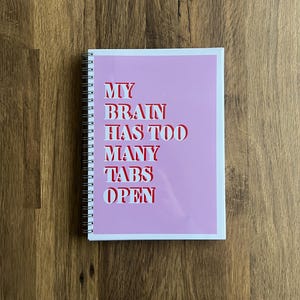 May include: A spiral-bound notebook with a pink cover. The cover features the text "MY BRAIN HAS TOO MANY TABS OPEN" in white and red lettering. The notebook is on a wooden surface.