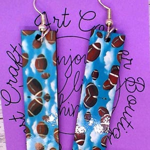 May include: A pair of rectangular earrings with a blue sky background and brown footballs. The earrings have silver-colored hooks. The background also has white clouds and silver accents. The earrings are approximately 2 inches long.
