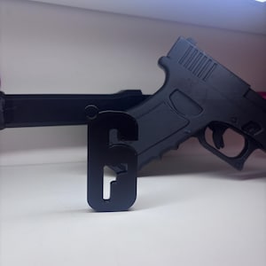 May include: Black metal gun display stand with the number 6 cut out. A black toy gun is resting on the stand. The stand is on a white surface.