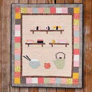 Puede incluir: Un tapiz de pared acolchado beige con un borde de patchwork colorido. El centro presenta una representación estilizada de un restaurante de sushi con estantes de rollos de sushi, un tazón de arroz y una tetera.