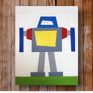 May include: A colorful, geometric robot design on a white background. The robot features a blue head, red arms, and a yellow torso with a polka dot pattern. Gray legs and feet stand on a green base. The artwork is displayed against a wooden backdrop.