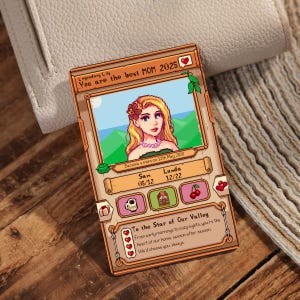 Może przedstawiać: Cyfrowa ilustracja kobiety z blond włosami i r&oacute;żowym kwiatem we włosach. Ilustracja jest w pikselowym stylu, podobnym do gry wideo Stardew Valley. Tekst głosi "Legendary Lily You are the best MOM 2025" i "Became a mom on 12th May 2020". Ilustracja jest otoczona brązową ramką z ikoną serca w prawym g&oacute;rnym rogu.