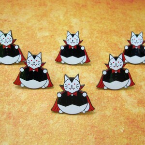 May include: Six enamel pins featuring a cartoon cat dressed as a vampire. The cat is white with black eyes and a black mouth. It is wearing a black cape with a red lining and a red bow tie. The cat is sitting on its haunches with its paws outstretched.