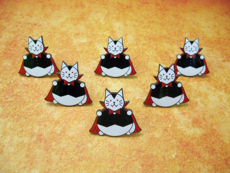 May include: Six enamel pins featuring a cartoon cat dressed as a vampire. The cat is white with black eyes and a black mouth. It is wearing a black cape with a red lining and a red bow tie. The cat is sitting on its haunches with its paws outstretched.