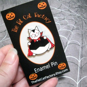 May include: A black and white enamel pin of a cat dressed as a vampire. The cat is wearing a red cape and has fangs. The pin is on a black card with the text "The Fat Cat Factory" and "Enamel Pin".