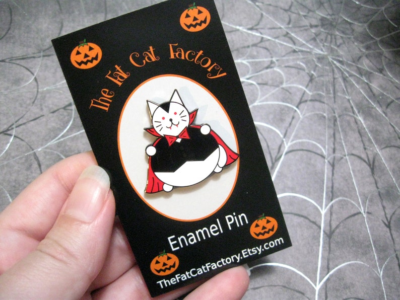 May include: A black and white enamel pin of a cat dressed as a vampire. The cat is wearing a red cape and has fangs. The pin is on a black card with the text "The Fat Cat Factory" and "Enamel Pin".