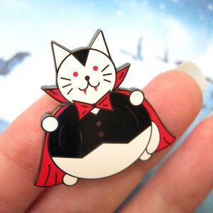 May include: A white cat dressed as a vampire with a red cape and black suit. The cat has red eyes and fangs. The cat is holding a red bow tie in its paws.