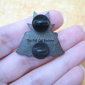 May include: Reverse of a silver enamel pin depicting a black cat wearing a cape. The pin has a black rubber clutch back and reads "The Fat Cat Factory" on the back.