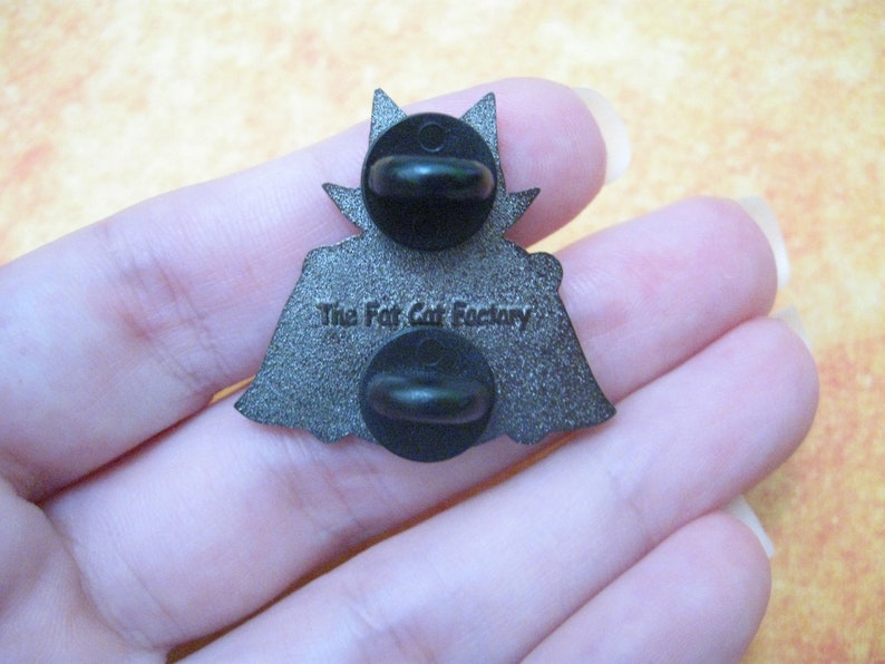 May include: Reverse of a silver enamel pin depicting a black cat wearing a cape. The pin has a black rubber clutch back and reads "The Fat Cat Factory" on the back.
