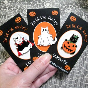 May include: Three enamel pins featuring Halloween-themed cats. The first pin shows a black cat dressed as a vampire, the second pin shows a white cat dressed as a ghost, and the third pin shows a black cat holding a jack-o-lantern. Each pin has the text "The Fat Cat Factory" on the card.