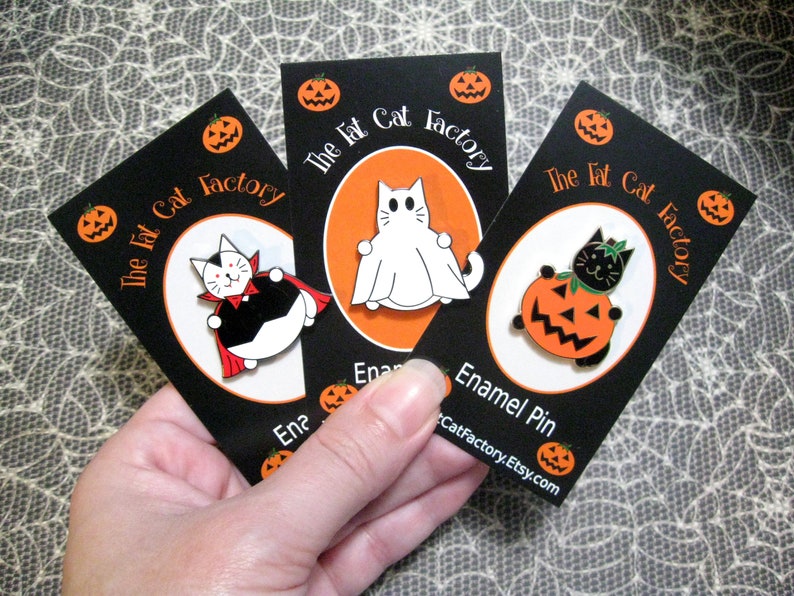 May include: Three enamel pins featuring Halloween-themed cats. The first pin shows a black cat dressed as a vampire, the second pin shows a white cat dressed as a ghost, and the third pin shows a black cat holding a jack-o-lantern. Each pin has the text "The Fat Cat Factory" on the card.