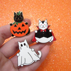 May include: Three enamel pins featuring black cats dressed up for Halloween. One cat is wearing a pumpkin costume, another is a vampire, and the third is a ghost.