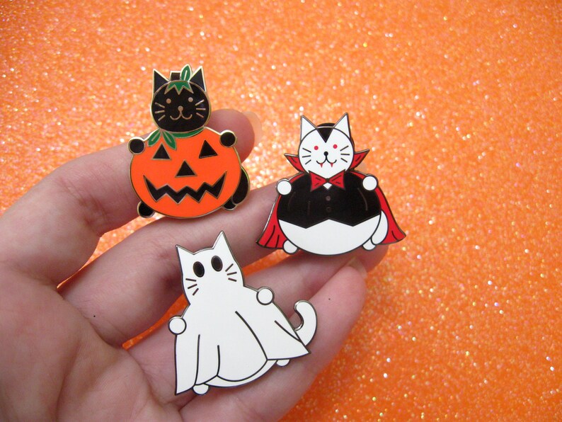 May include: Three enamel pins featuring black cats dressed up for Halloween. One cat is wearing a pumpkin costume, another is a vampire, and the third is a ghost.