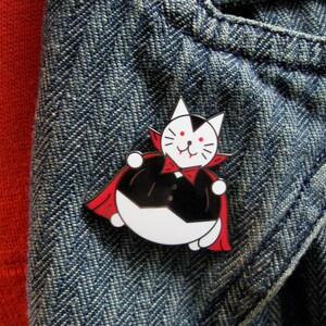 May include: A white cat wearing a red cape and a black tuxedo, with fangs, is depicted as a vampire. The cat is a pin on a blue denim background.