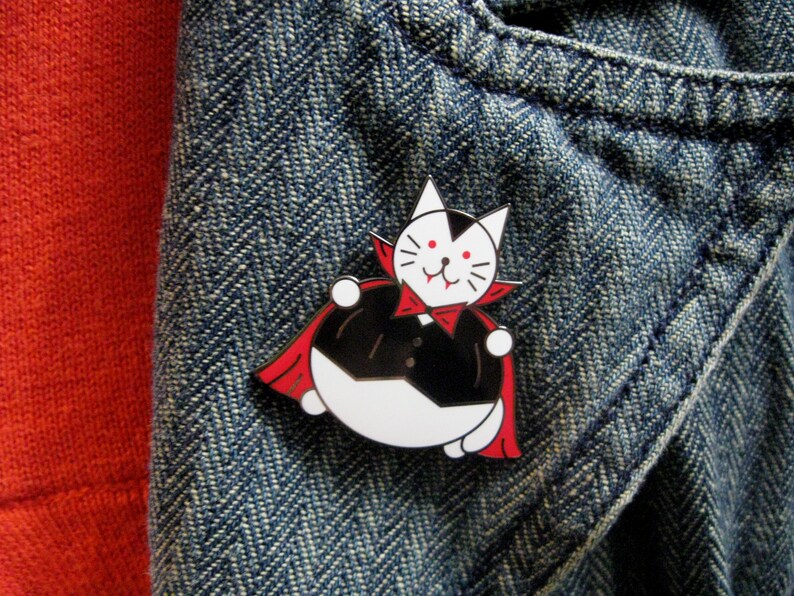 May include: A white cat wearing a red cape and a black tuxedo, with fangs, is depicted as a vampire. The cat is a pin on a blue denim background.