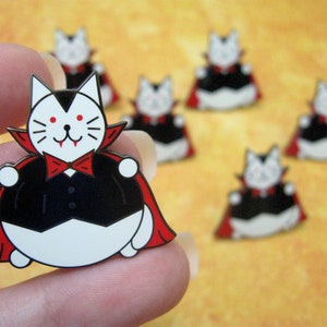 May include: A set of seven enamel pins featuring a cartoon cat dressed as a vampire. The cat is wearing a black cape with a red lining, a black bow tie, and a white collar. The cat has red eyes and fangs. The pins are a fun and festive way to add a touch of Halloween to your clothing or accessories.