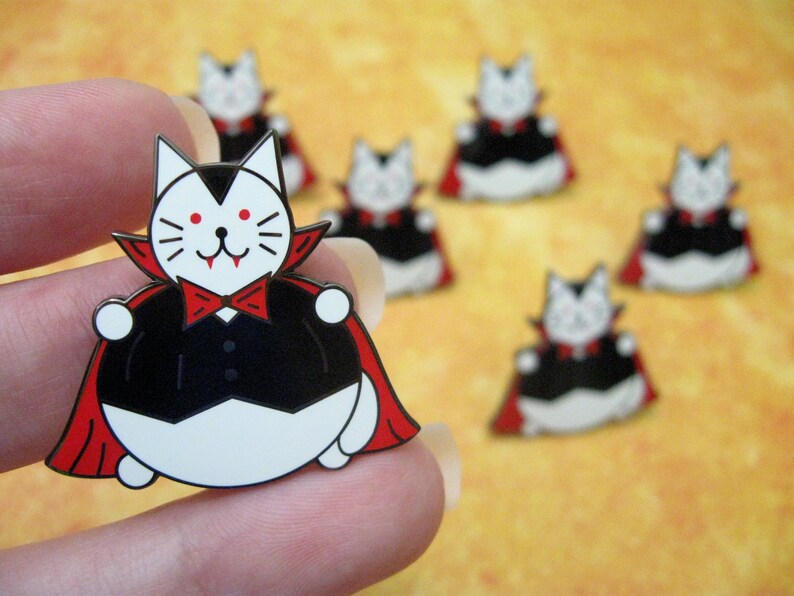 May include: A set of seven enamel pins featuring a cartoon cat dressed as a vampire. The cat is wearing a black cape with a red lining, a black bow tie, and a white collar. The cat has red eyes and fangs. The pins are a fun and festive way to add a touch of Halloween to your clothing or accessories.