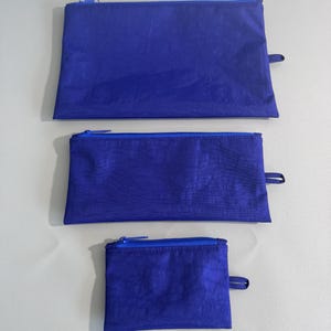 May include: Three blue zippered pouches of varying sizes. The pouches are made of a textured material and have a matching blue zipper and a small fabric loop on one end. The pouches are arranged from largest to smallest.
