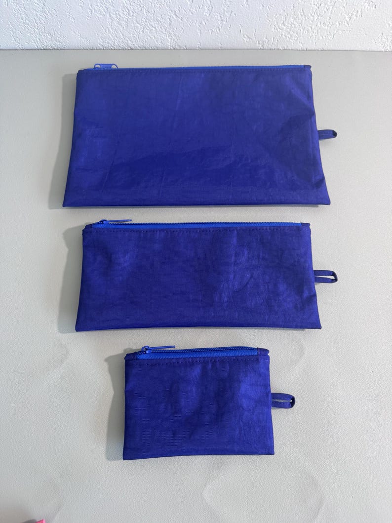 May include: Three blue zippered pouches of varying sizes. The pouches are made of a textured material and have a matching blue zipper and a small fabric loop on one end. The pouches are arranged from largest to smallest.