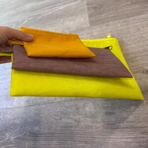 May include: Three colorful pouches in shades of yellow, brown, and orange. The pouches are stacked on top of each other, with the smallest orange pouch on top. The pouches are made of a soft, flexible material.