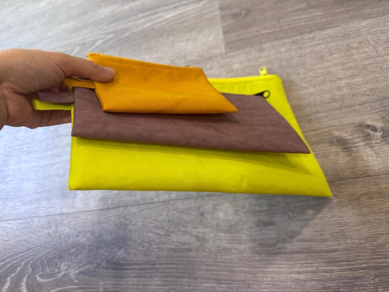 May include: Three colorful pouches in shades of yellow, brown, and orange. The pouches are stacked on top of each other, with the smallest orange pouch on top. The pouches are made of a soft, flexible material.