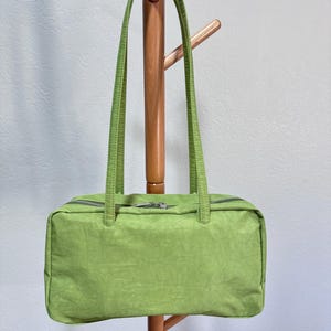 May include: A rectangular, lime green handbag with two long, matching handles. The bag has a zip closure and is hanging on a wooden coat rack. The bag's material has a slightly textured appearance.