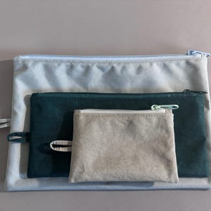 May include: Three zippered pouches in varying sizes and colors. The largest is a light gray, the middle one is dark teal, and the smallest is a light beige. Each pouch has a matching zipper and a small fabric loop.