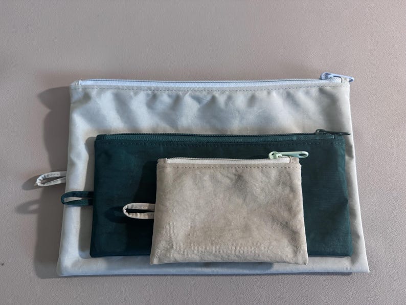 May include: Three zippered pouches in varying sizes and colors. The largest is a light gray, the middle one is dark teal, and the smallest is a light beige. Each pouch has a matching zipper and a small fabric loop.