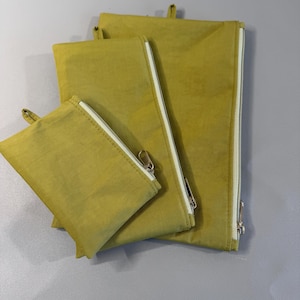 May include: Four olive-green zippered pouches of varying sizes. Each pouch has a white zipper and a small loop at the top. The pouches are made of a soft, textured fabric and are arranged in a nested configuration.