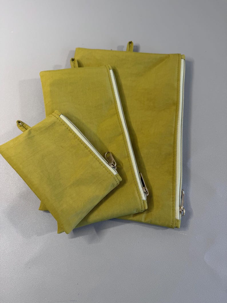 May include: Four olive-green zippered pouches of varying sizes. Each pouch has a white zipper and a small loop at the top. The pouches are made of a soft, textured fabric and are arranged in a nested configuration.