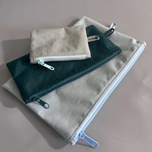 May include: A set of four zippered pouches in varying sizes and colors. The smallest pouch is a light beige, the next is a deep teal, and the two largest are a light gray. Each pouch has a matching zipper.