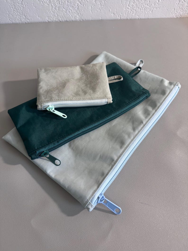 May include: A set of four zippered pouches in varying sizes and colors. The smallest pouch is a light beige, the next is a deep teal, and the two largest are a light gray. Each pouch has a matching zipper.