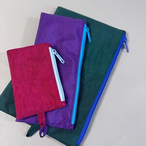 May include: Three colorful zippered pouches in varying sizes. The smallest is a deep magenta, the middle one is purple, and the largest is a dark teal. Each pouch has a contrasting zipper color: light blue, light blue, and blue.