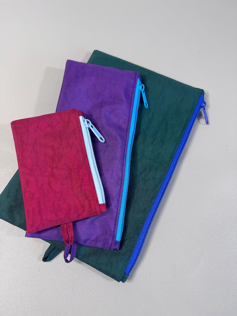 May include: Three colorful zippered pouches in varying sizes. The smallest is a deep magenta, the middle one is purple, and the largest is a dark teal. Each pouch has a contrasting zipper color: light blue, light blue, and blue.
