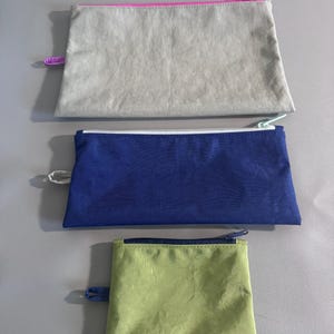 May include: Three rectangular zippered pouches in varying sizes. The top pouch is light gray with a pink zipper. The middle pouch is navy blue with a white zipper. The bottom pouch is light green with a dark blue zipper.