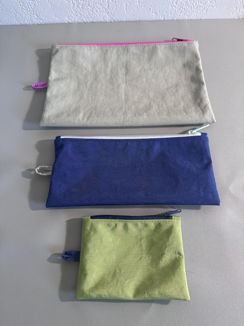 May include: Three rectangular zippered pouches in varying sizes. The top pouch is light gray with a pink zipper. The middle pouch is navy blue with a white zipper. The bottom pouch is light green with a dark blue zipper.