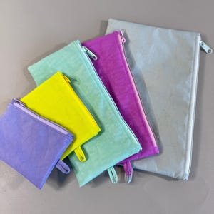 May include: A collection of colorful zippered pouches in various sizes. The pouches are in shades of purple, yellow, teal, and gray. Each pouch has a matching zipper and pull tab.